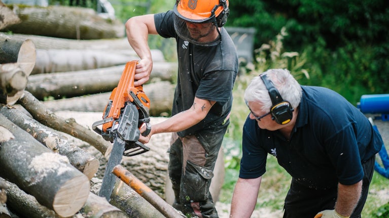Ranger in a black t-shirt with the National Trust oak leaf white logo, wearing an orange safety hat and using a chainsaw. Next to him is a volunteer, wearing ear defenders, who is helping by holding the wood steady.
